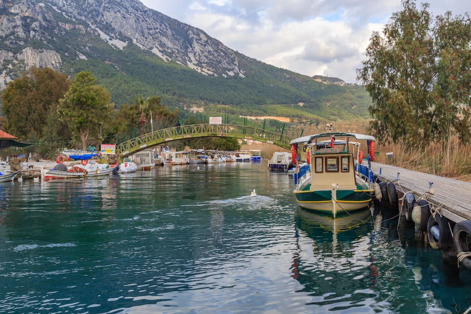 Un río con barcos amarrados, un puente de madera y un paisaje montañoso al fondo.