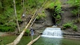 Ein Wasserfall in bewaldeter Landschaft, eine Person steht auf einem Baumstamm.