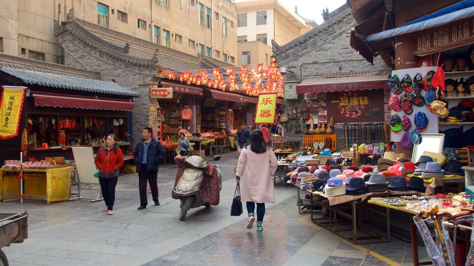 A bustling market street with traditional architecture, various stalls selling hats and accessories, and people walking through the area.