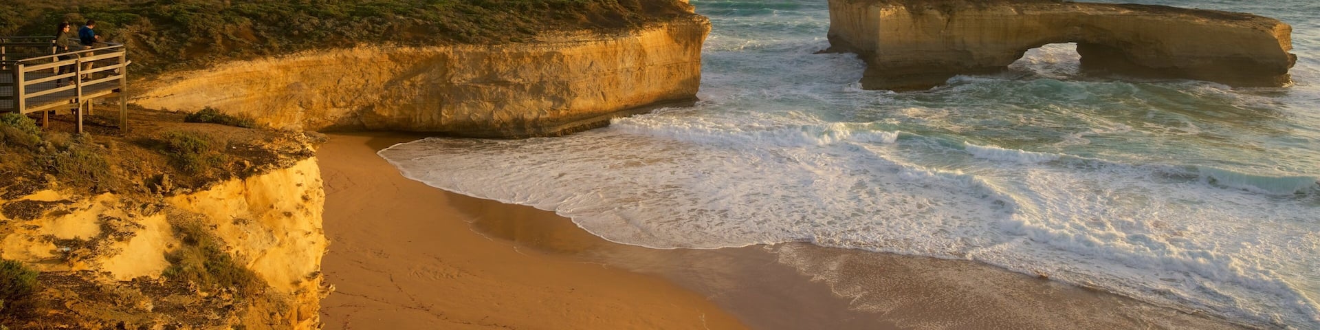 A coastal landscape with a sandy beach, a wooden viewing platform, and a large rock formation in the sea.