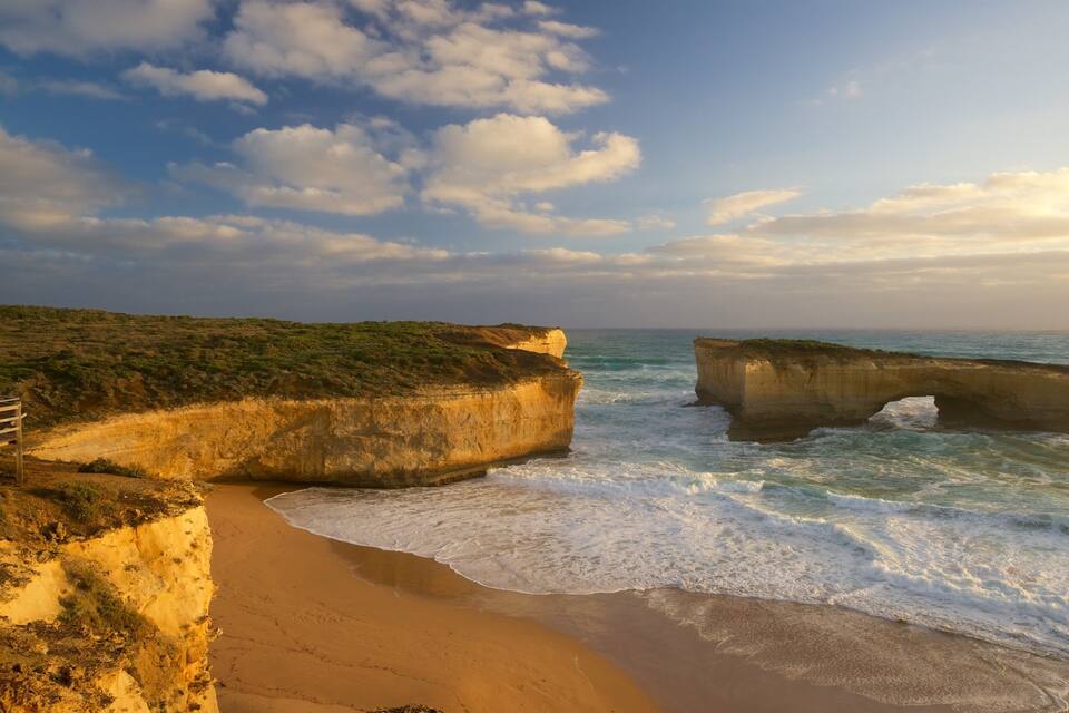 A coastal landscape with a sandy beach, a wooden viewing platform, and a large rock formation in the sea.