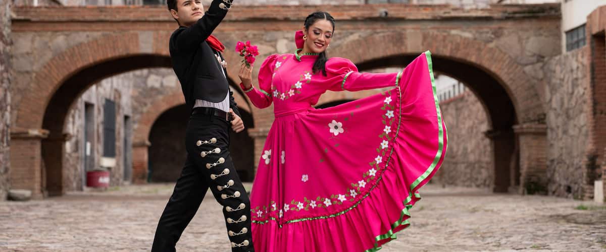 A couple in traditional Mexican attire dancing in an old stone courtyard with arches.