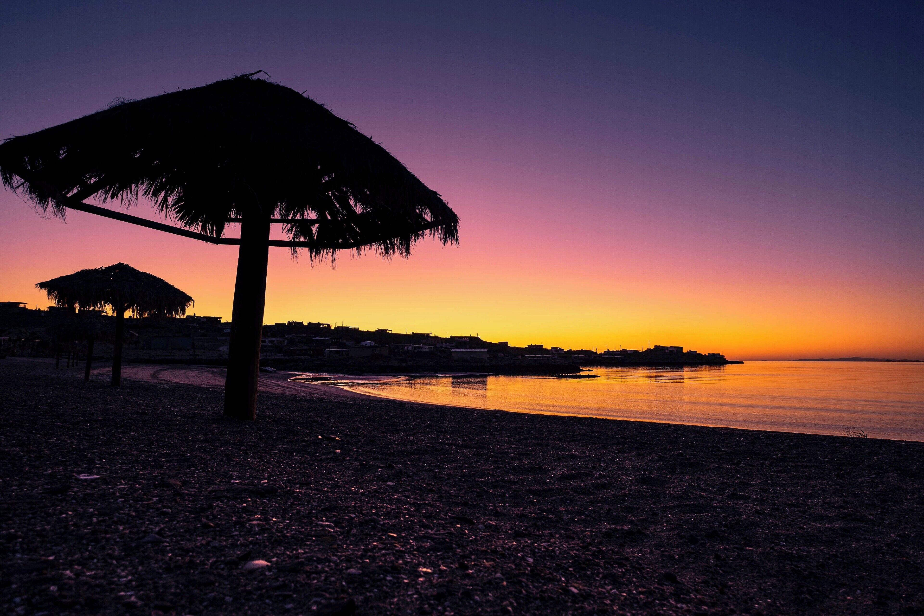 A beach at sunset with a thatched umbrella and a calm sea.