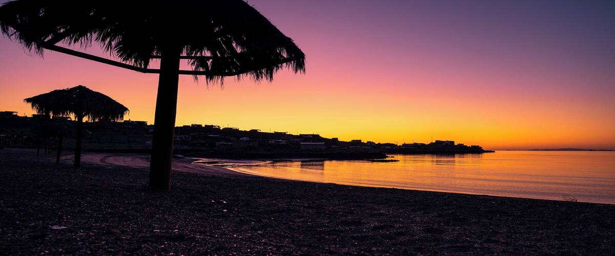 A beach at sunset with a thatched umbrella and a calm sea.