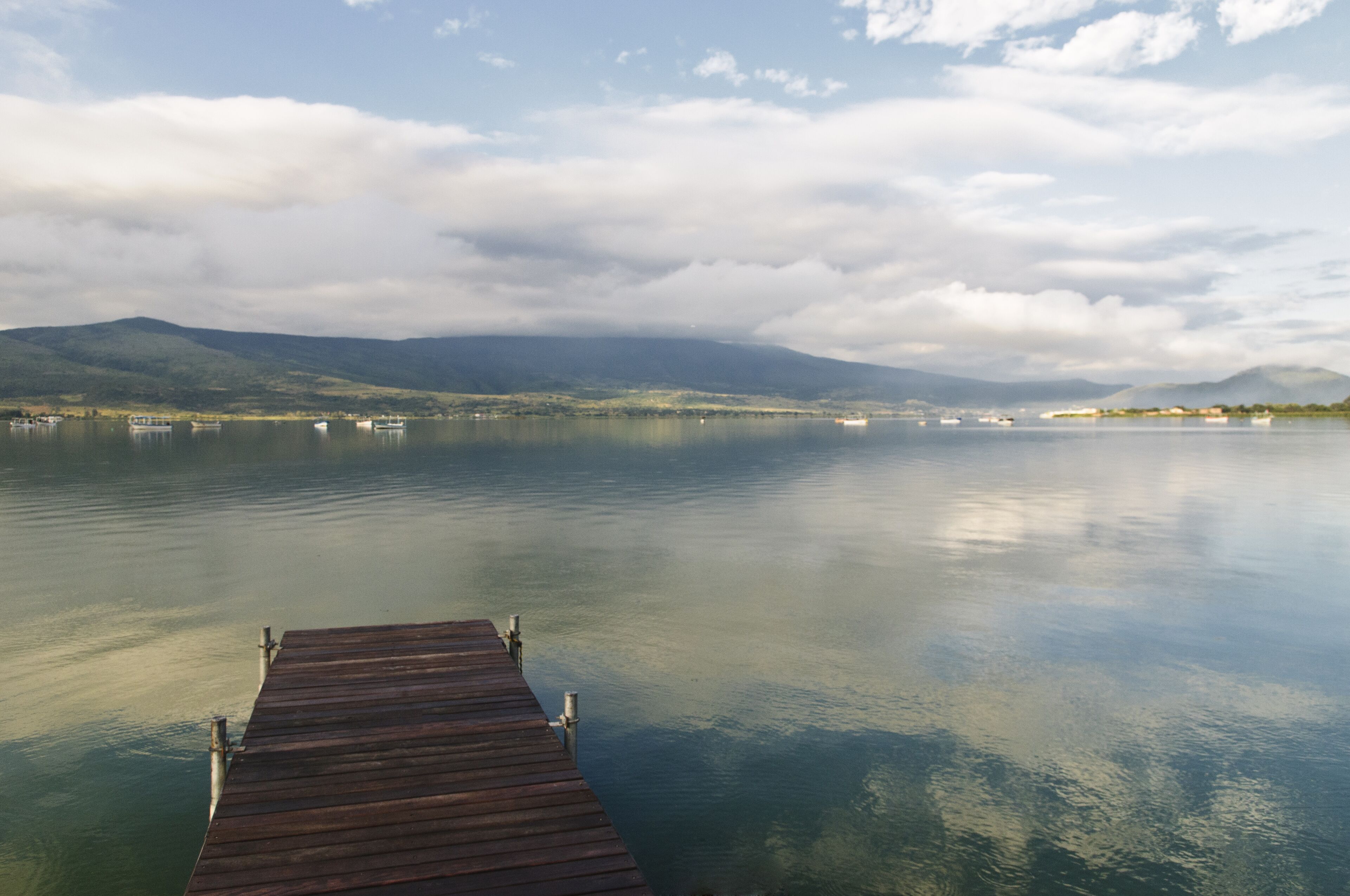 A wooden pier extending into calm waters with mountains in the background and a partly cloudy sky.
