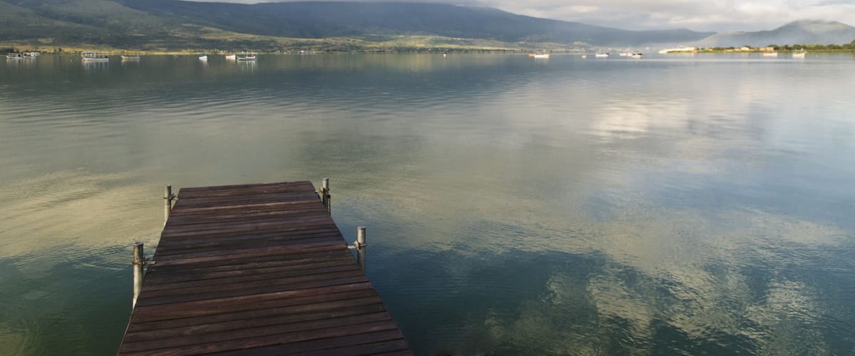 A wooden pier extending into calm waters with mountains in the background and a partly cloudy sky.