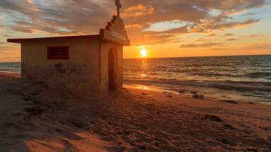 A small building with a cross on top, located on a beach with the ocean in the background and the sun setting.