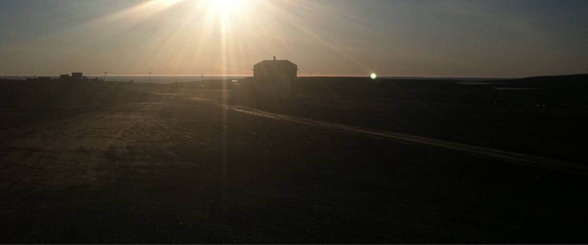 A bright sun low on the horizon, casting a glow over a flat landscape with a solitary building in the distance.