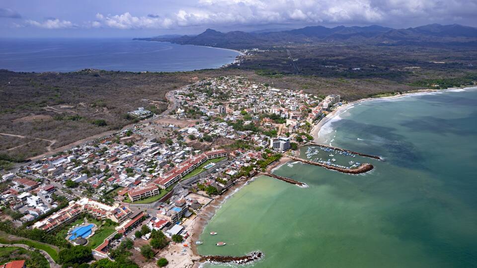 An aerial view of a coastal town with a beach, marina, and mountainous background.