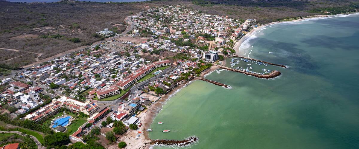 An aerial view of a coastal town with a beach, marina, and mountainous background.
