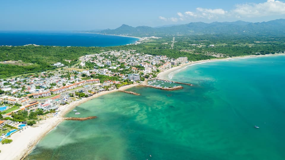 An aerial view of a coastal town with a beach, clear turquoise waters, and a mountainous backdrop.