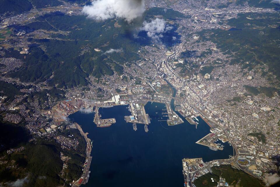 Vista aerea di una città costiera con un ampio porto, aree urbane fitte e colline verdi circostanti.
