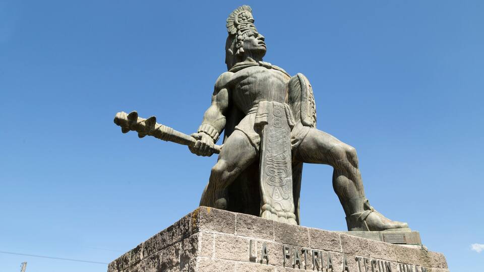 A statue of a seated figure with a spear, wearing a headdress, and inscribed with "LA PATRIA A TECUN UMAN".