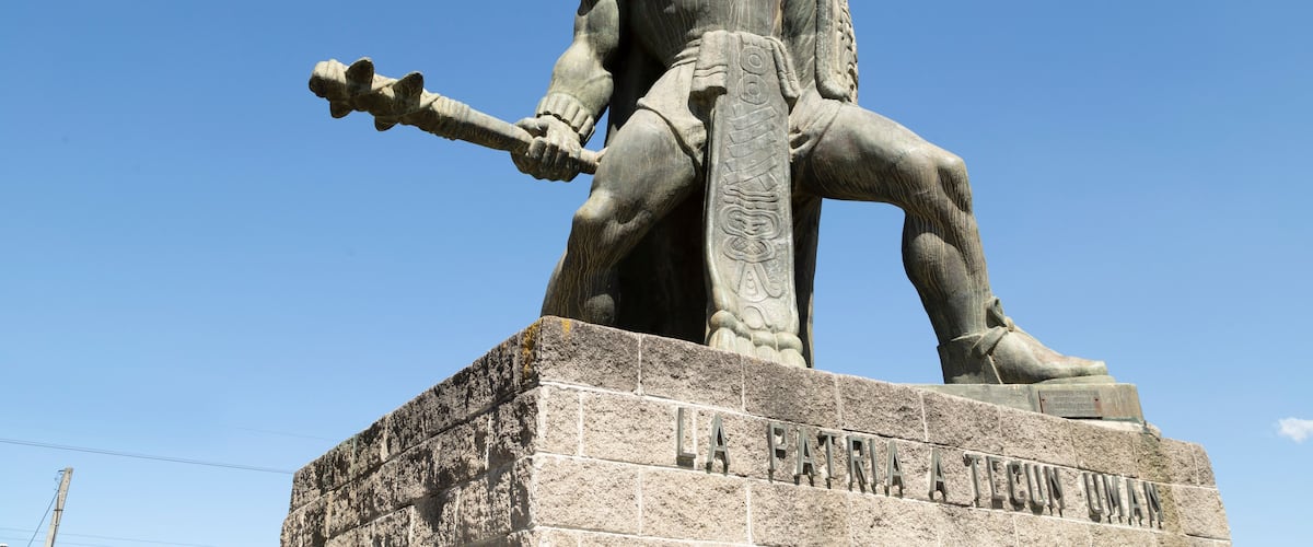 A statue of a seated figure with a spear, wearing a headdress, and inscribed with "LA PATRIA A TECUN UMAN".