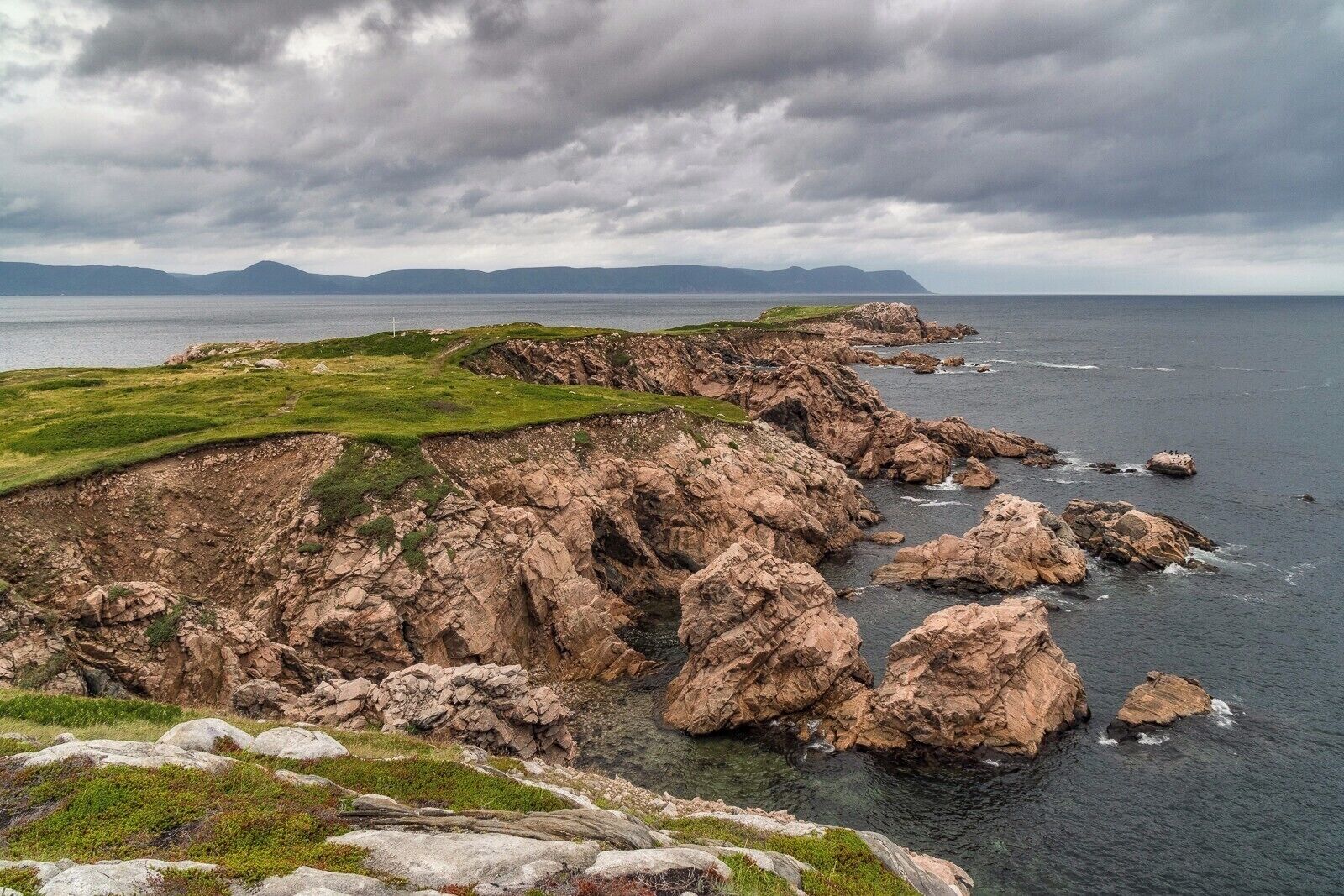 A rugged coastline with rocky cliffs, grassy areas, and a distant view of mountains under a cloudy sky.