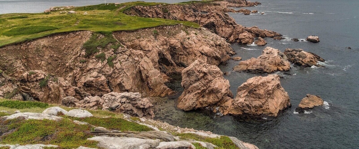 A rugged coastline with rocky cliffs, grassy areas, and a distant view of mountains under a cloudy sky.