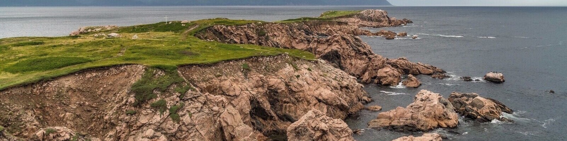 A rugged coastline with rocky cliffs, grassy areas, and a distant view of mountains under a cloudy sky.