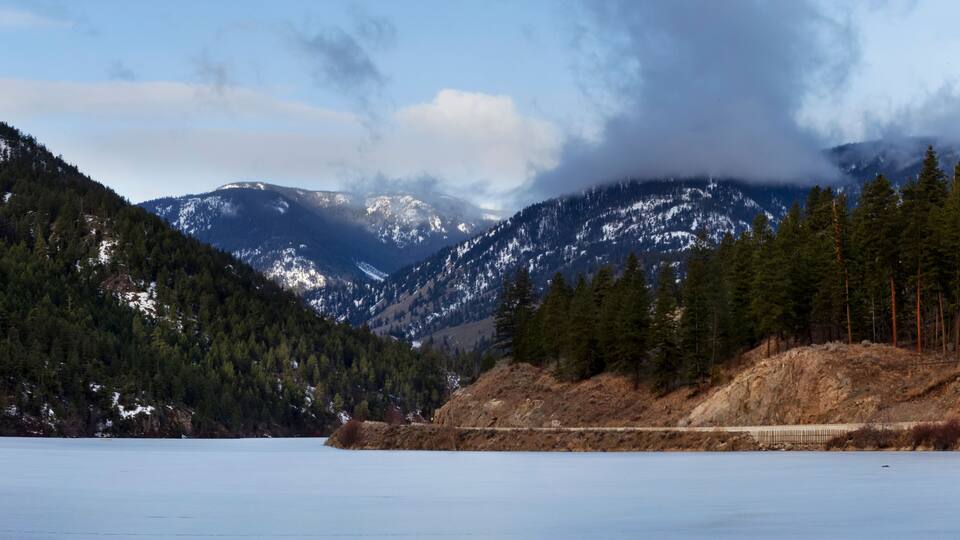 A frozen lake with snow-capped mountains in the background and a forested hillside.