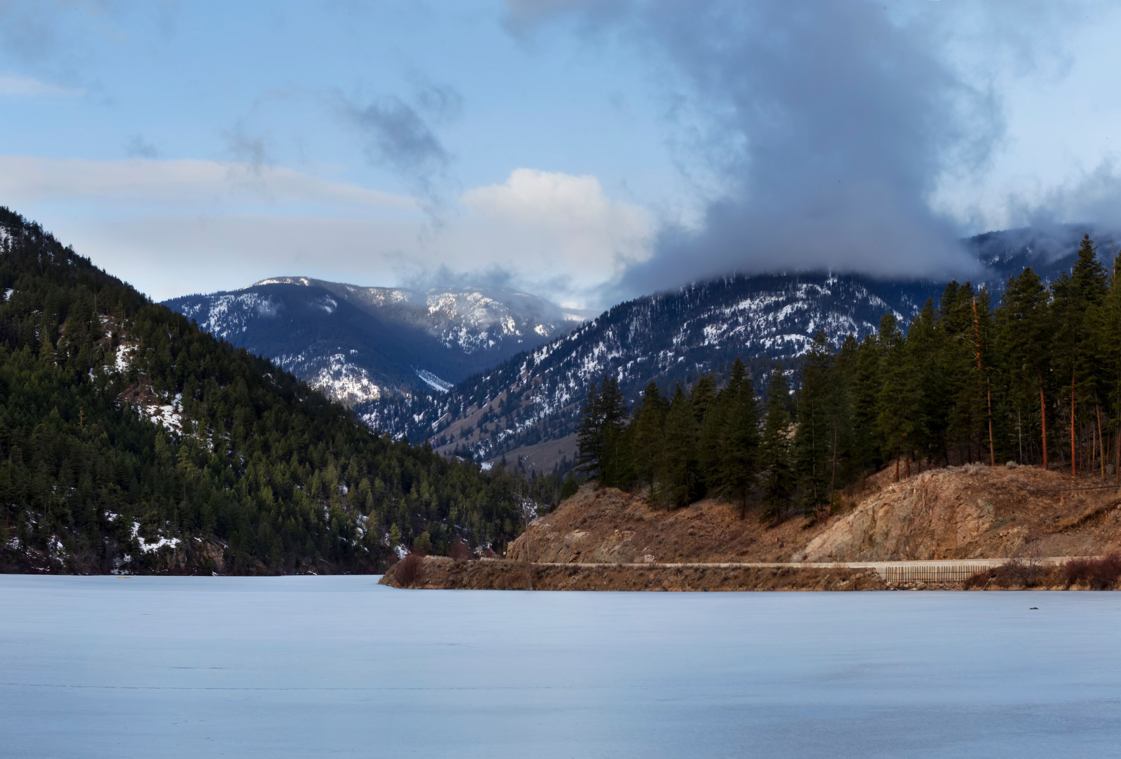 A frozen lake with snow-capped mountains in the background and a forested hillside.