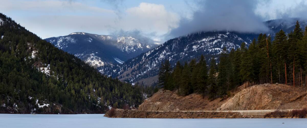 A frozen lake with snow-capped mountains in the background and a forested hillside.