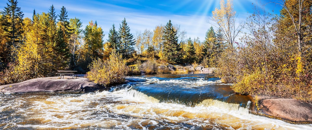 A river with rapids, surrounded by autumn-colored trees and a bench.