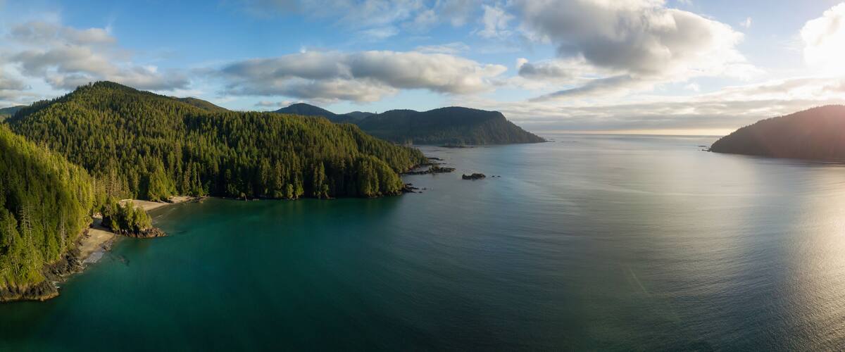 A coastal landscape with dense forests, a calm sea, and distant mountains under a partly cloudy sky.