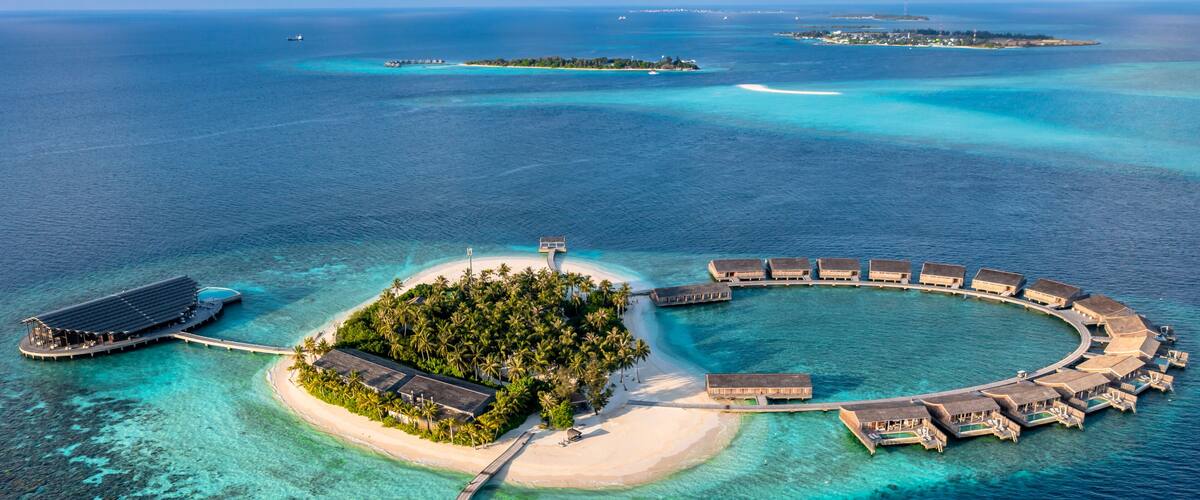 An aerial view of a tropical island resort with overwater bungalows, a central building, and clear turquoise waters.