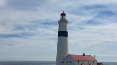 A lighthouse with a red roof and white walls, next to a small white building with a red roof, set against a cloudy sky.