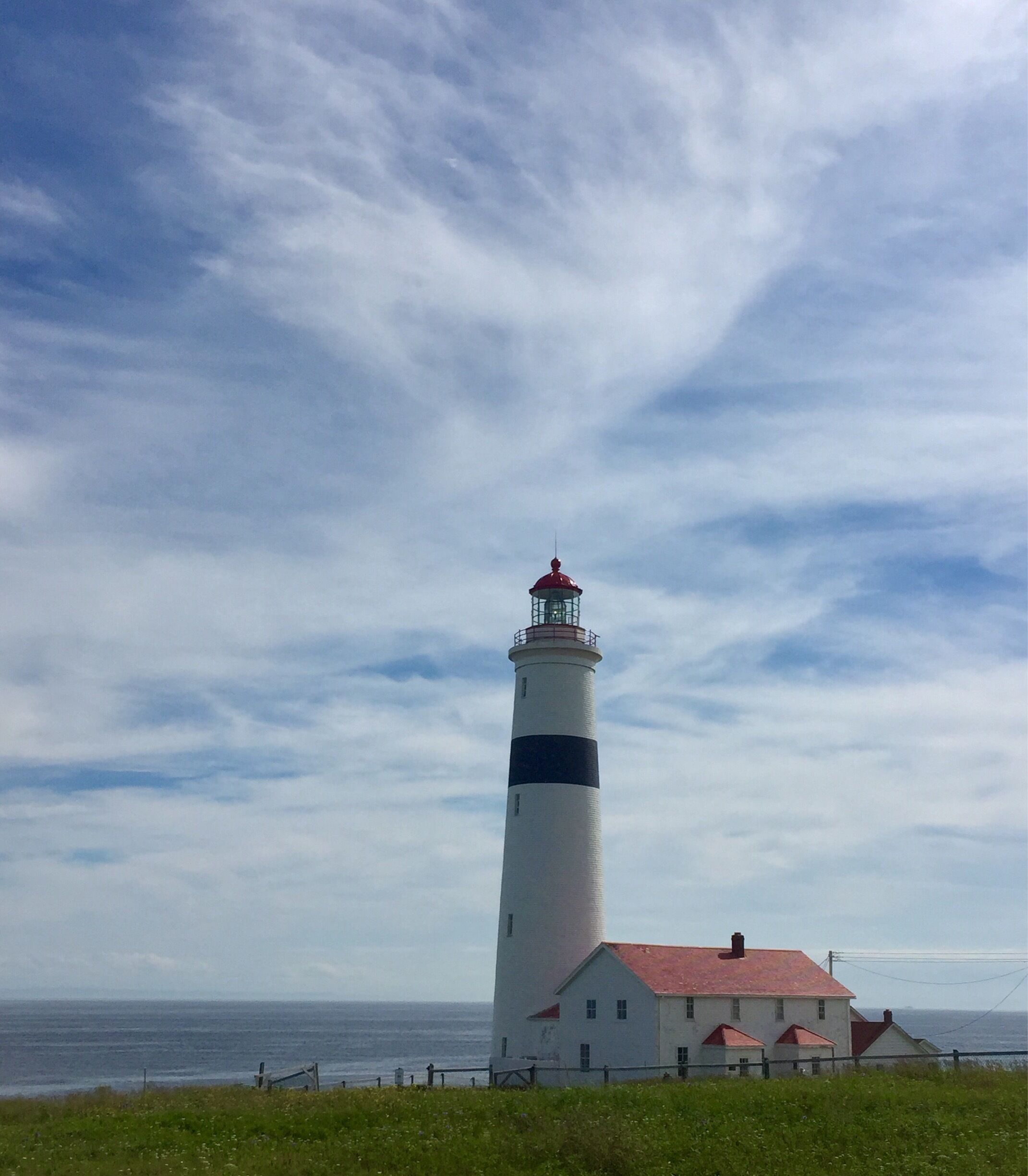 A lighthouse with a red roof and white walls, next to a small white building with a red roof, set against a cloudy sky.