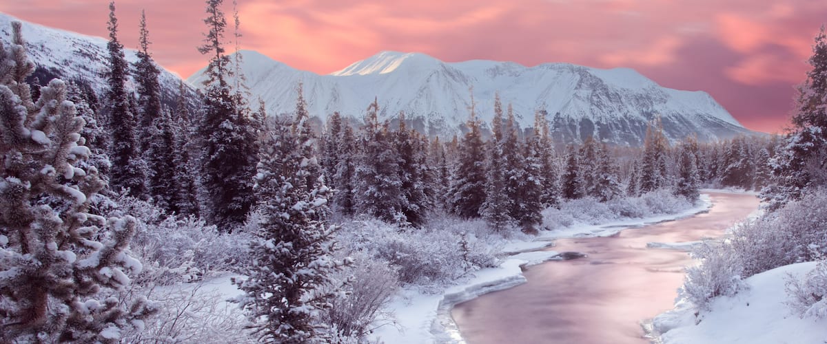 A snow-covered river winding through a forest with mountains in the background.