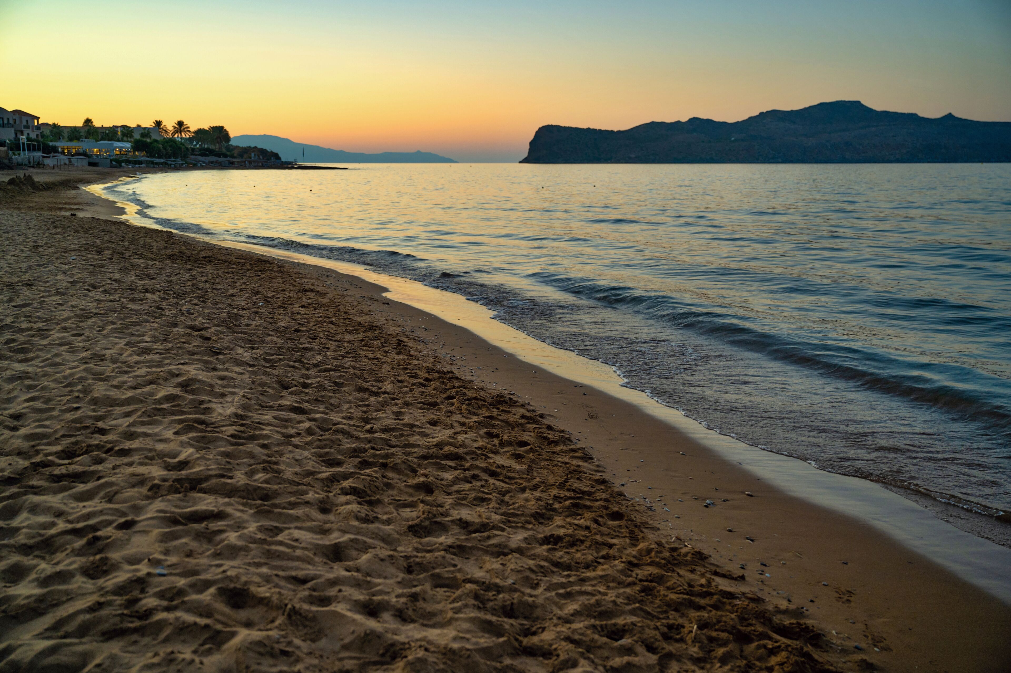 A beach with sand and water, a distant mountain, and a sunset.
