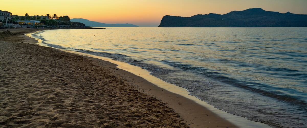 A beach with sand and water, a distant mountain, and a sunset.