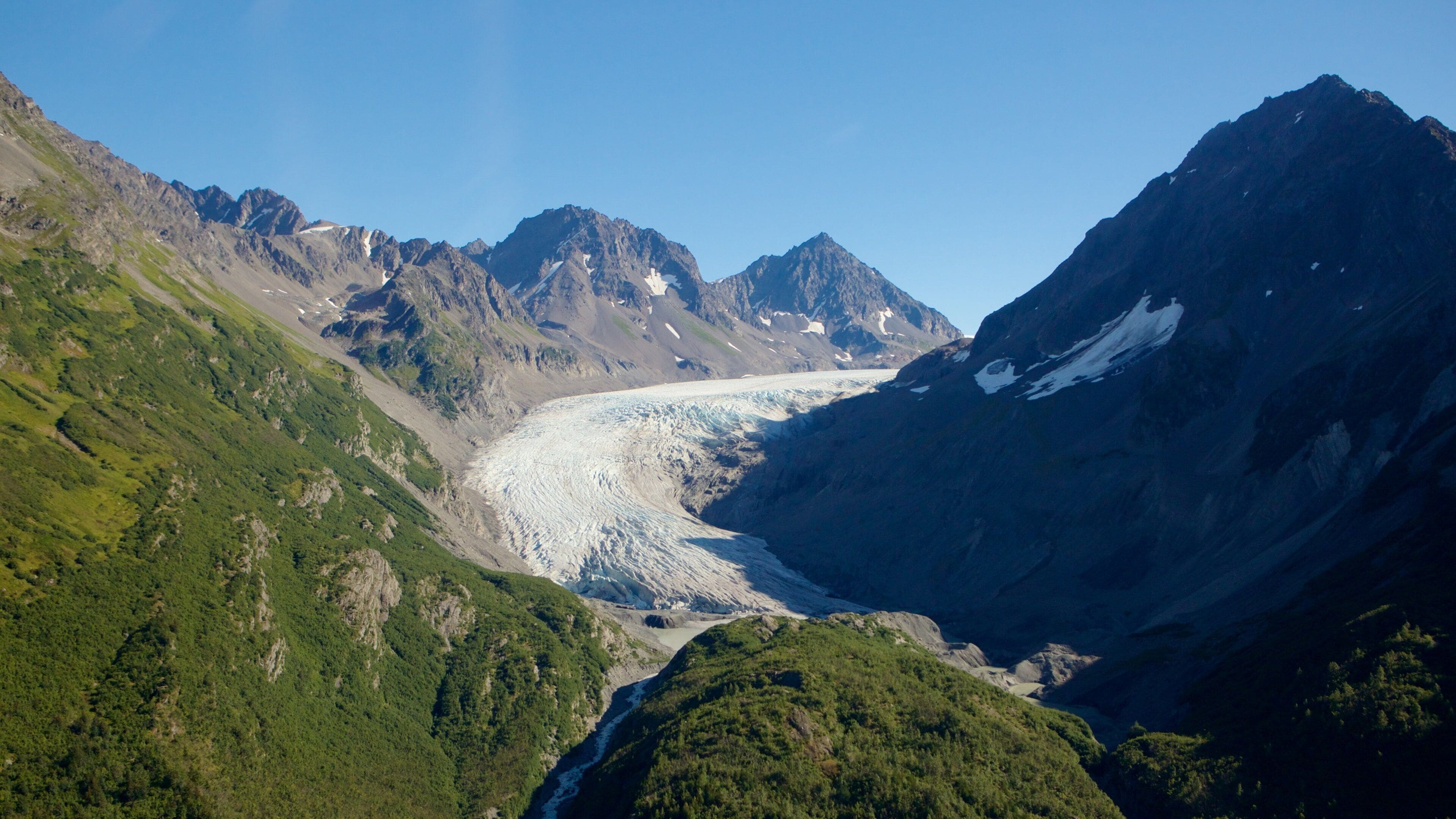 Um glaciar em um vale montanhoso, com picos rochosos e vegetação exuberante.