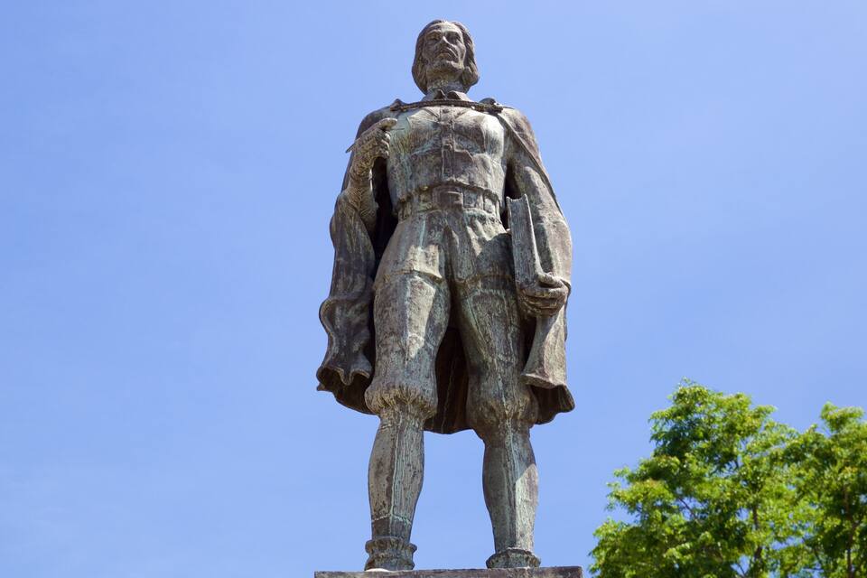 A statue of a man in historical attire, standing on a pedestal with a clear blue sky and a tree in the background.