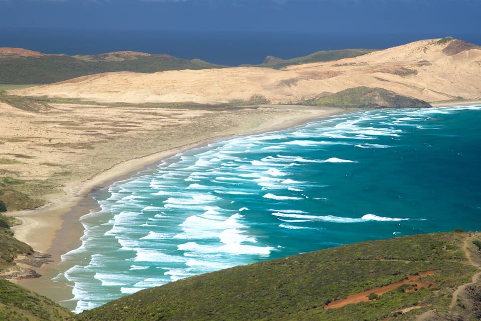 A coastal landscape with a sandy beach, turquoise ocean waves, and green hills.