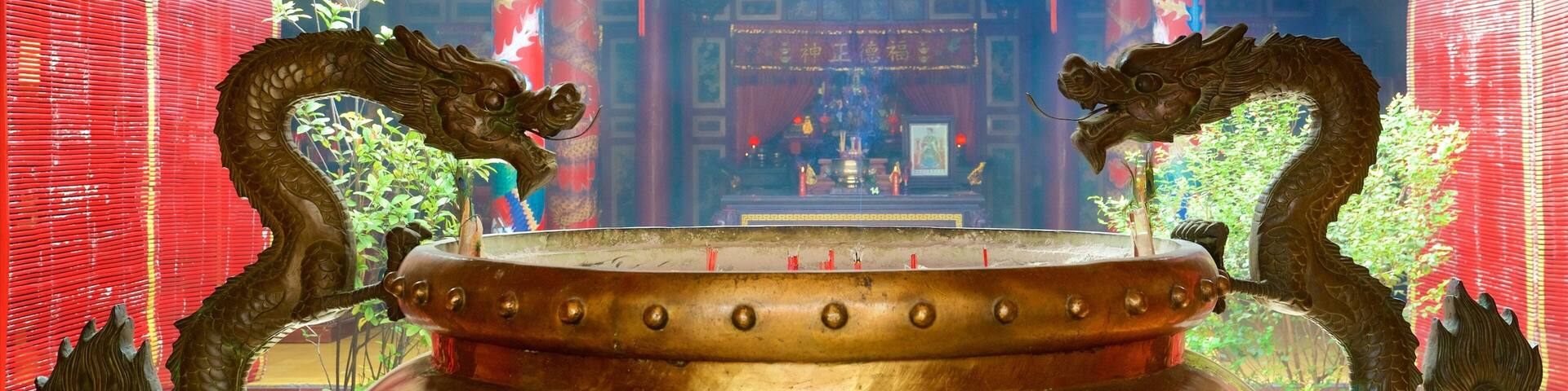 A large bronze incense burner with dragon handles, set against a temple backdrop with red walls and traditional Chinese decorations.
