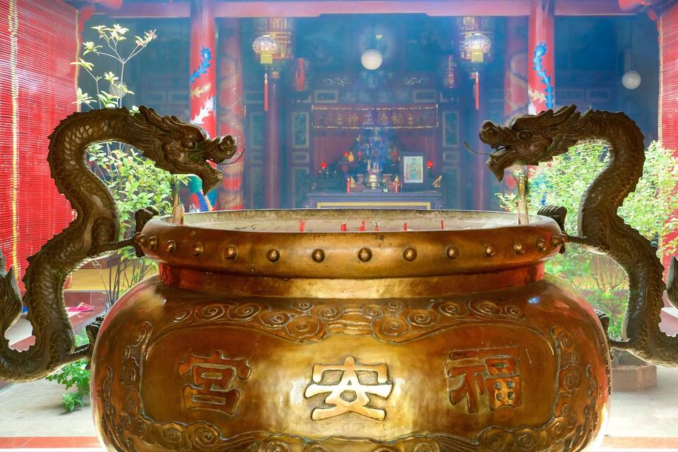 Un ampio bruciatore di incenso in bronzo con manici a forma di drago, sullo sfondo di un tempio con pareti rosse e decorazioni tradizionali cinesi.