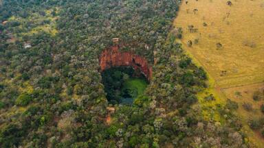 Vista aérea de uma área arborizada com uma grande formação rochosa vermelha, cercada por árvores e um campo à direita.