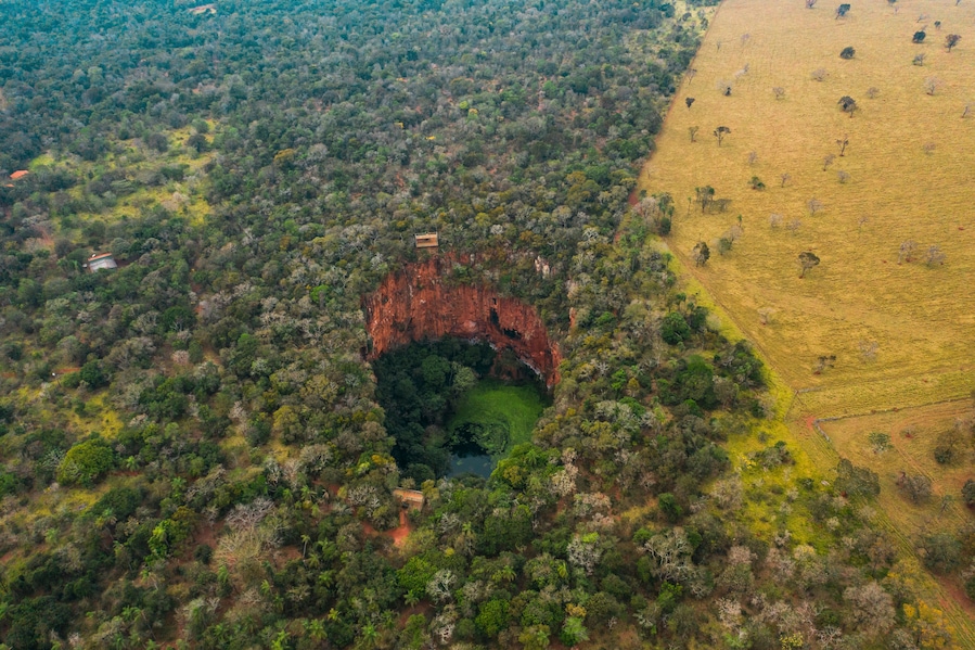 Vista aérea de uma área arborizada com uma grande formação rochosa vermelha, cercada por árvores e um campo à direita.