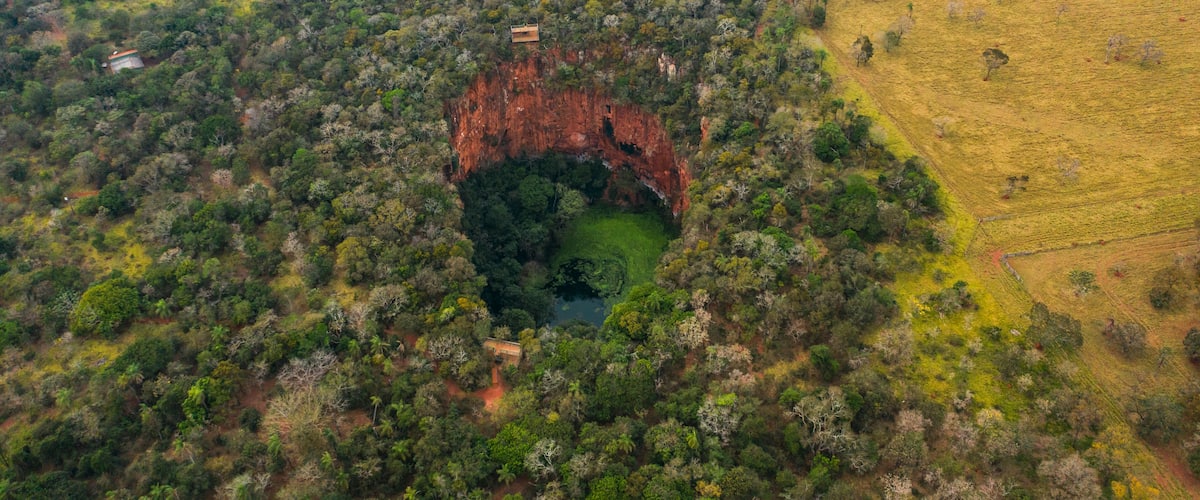 An aerial view of a forested area with a large red rock formation surrounded by trees and a field on the right.