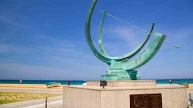 A large, curved metal sculpture with inscriptions, located near a beach with a clear blue sky.