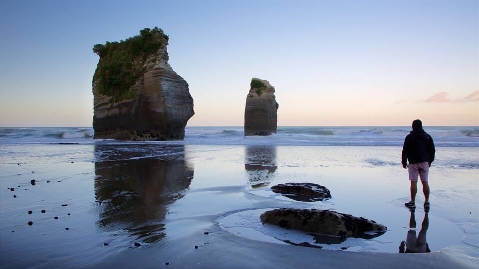 A person standing on a beach with large rock formations in the background.