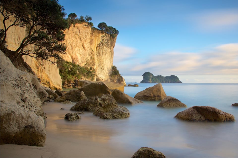 A coastal landscape with large boulders, a sandy beach, and a cliff with sparse vegetation.
