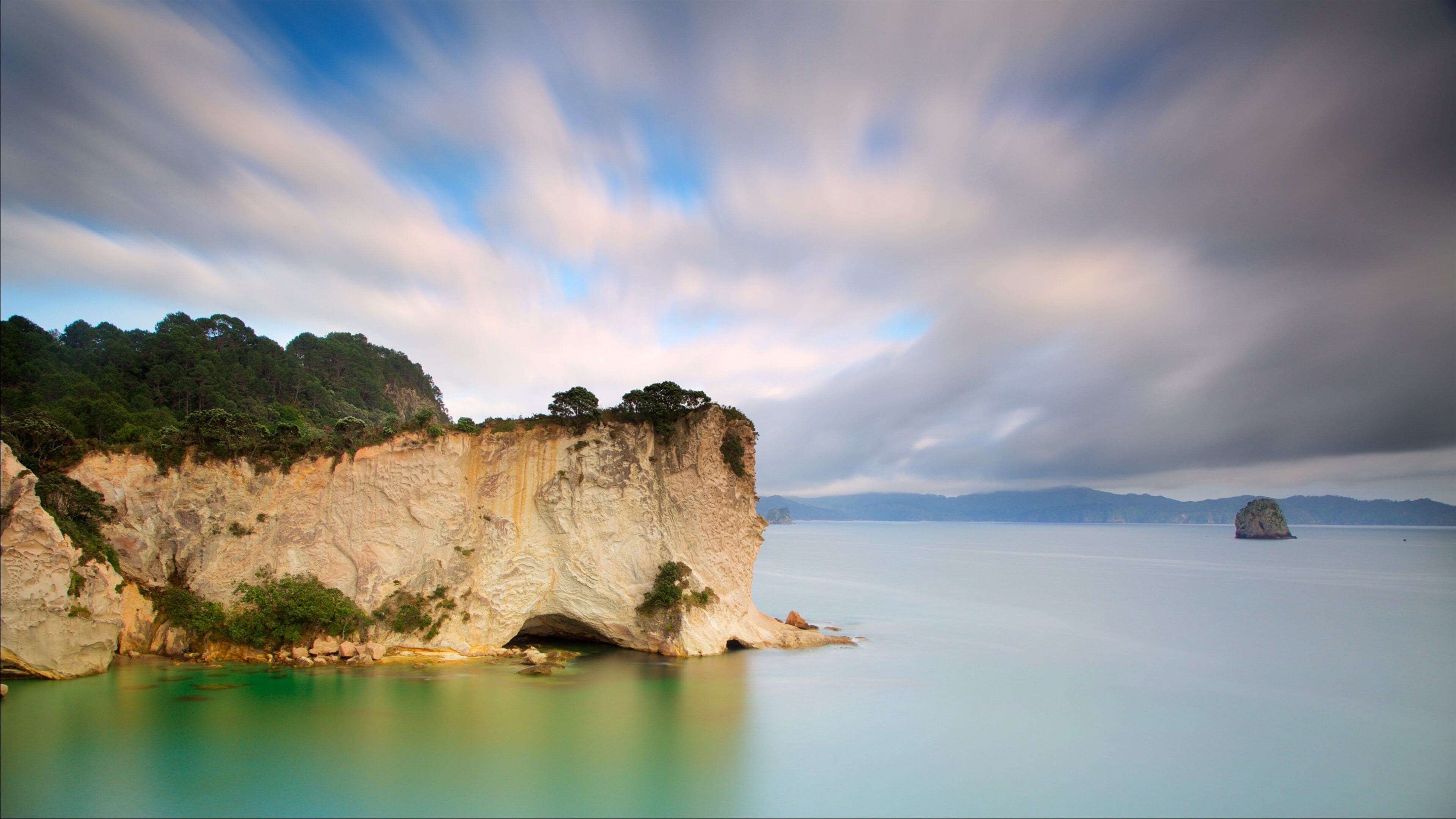 A coastal cliff with a cave entrance, calm turquoise waters, and distant mountains under a cloudy sky.