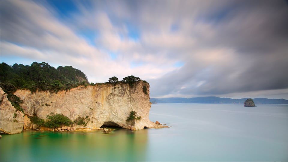 A coastal cliff with a cave entrance, calm turquoise waters, and distant mountains under a cloudy sky.