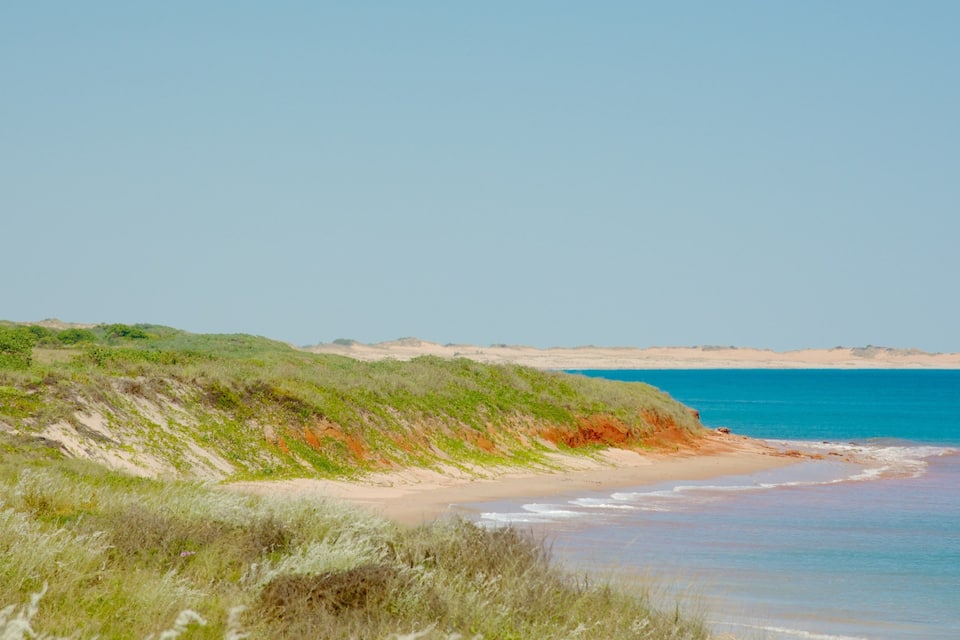 A coastal landscape with a sandy beach, grassy dunes, and a clear blue sea.