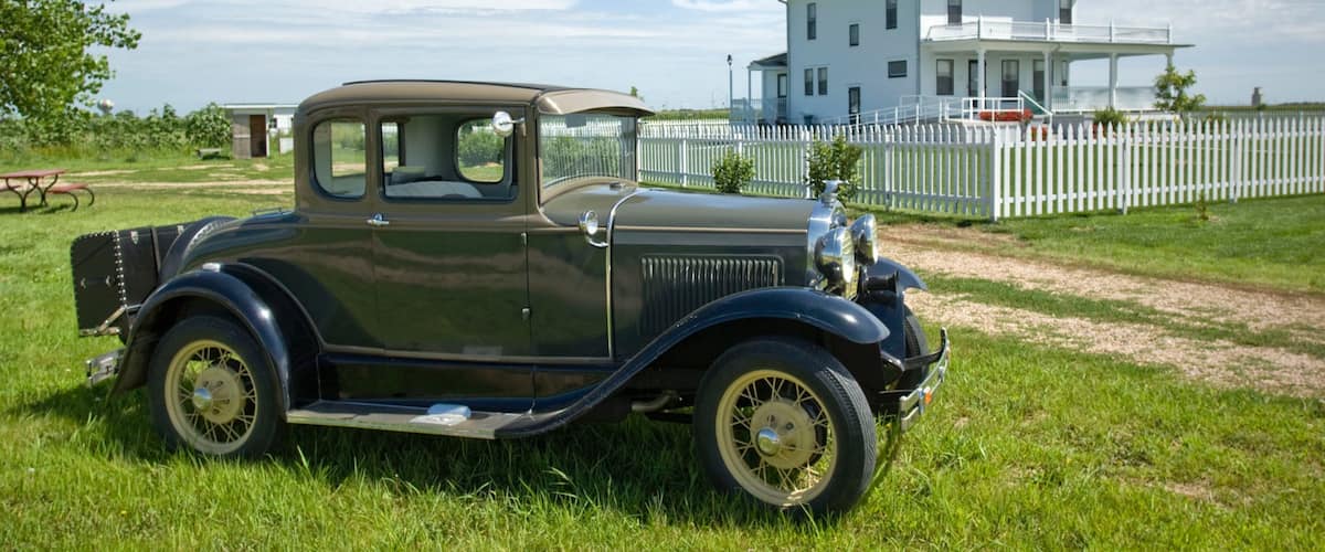 A vintage car parked on grass with a white house and white picket fence in the background.