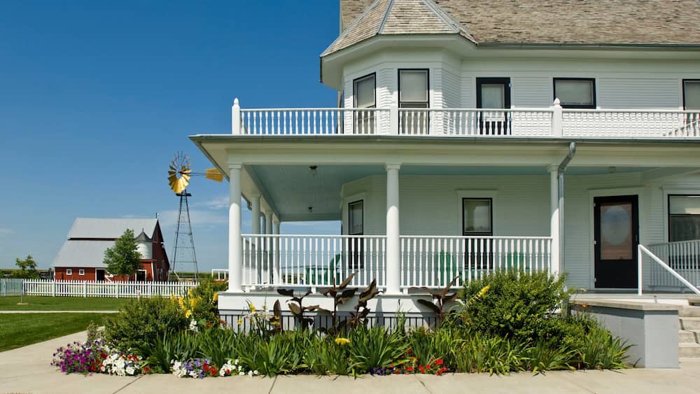 A white two-story house with a balcony, surrounded by a garden with various flowers.