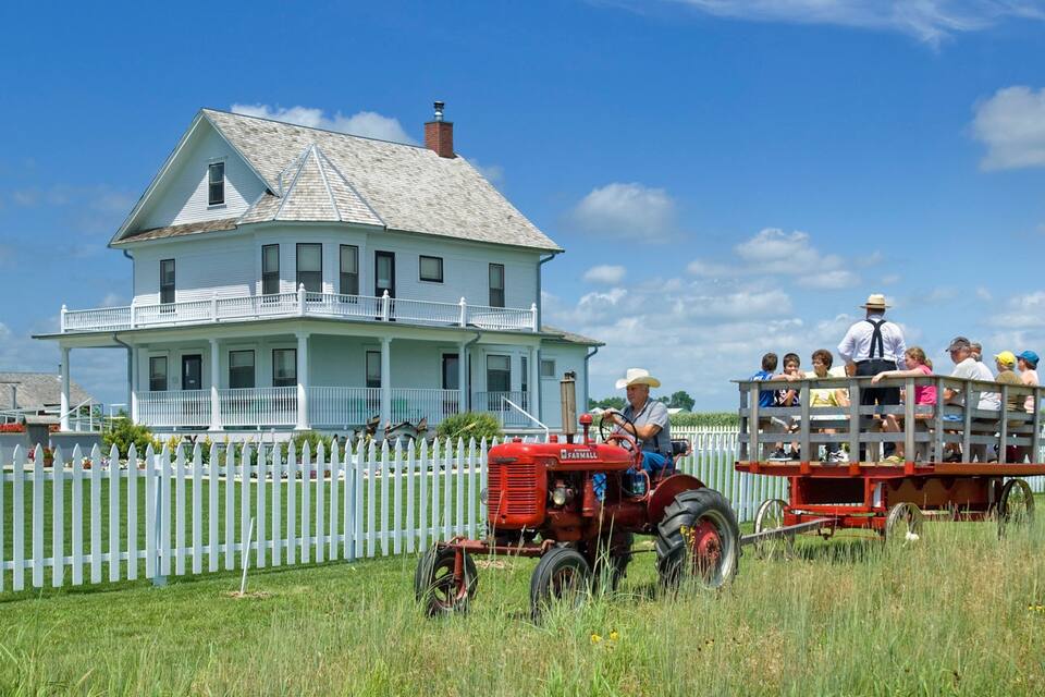Un tracteur rouge tirant une charrette remplie de personnes, une maison blanche avec un porche et une clôture blanche en piquets.