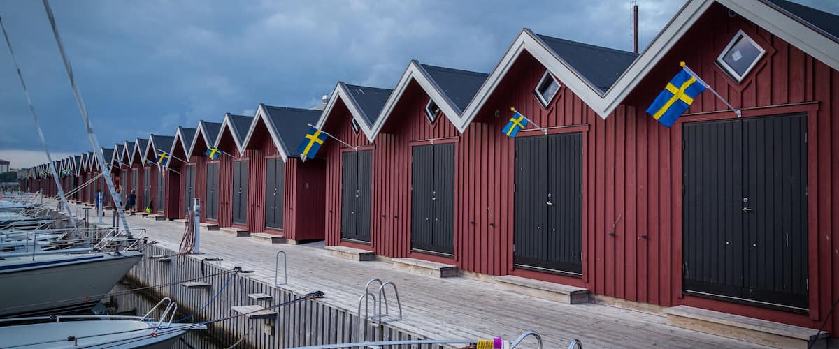 A row of red wooden buildings with Swedish flags, a wooden dock, and boats.